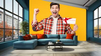 Young happy man he wears red checkered shirt casual clothes hold gift certificate coupon voucher card for store do winner gesture isolated on plain yellow orange background studio. Lifestyle concept. Wall mural