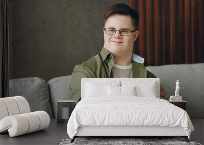 Young happy man with down syndrome wear glasses casual clothes use mobile cell phone sits on grey sofa couch stay at home flat rest spend free time in living room. Genetic disease world day concept. Wall mural