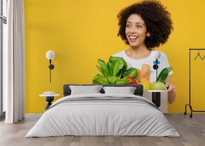 Young happy smiling amazed woman of African American ethnicity wears white t-shirt hold box with vegetarian vegan food organic vegetables look aside on workspace isolated on plain yellow background Wall mural