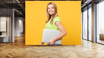 Young happy smiling cheerful blonde IT woman wear green t-shirt casual clothes hold closed laptop pc computer look camera isolated on plain yellow orange background studio portrait. Lifestyle concept. Wall mural