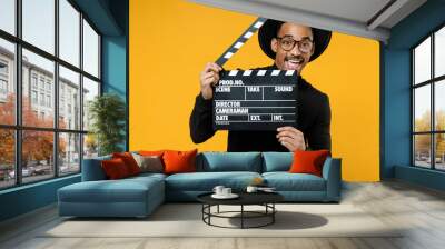 Young happy smiling fun cheerful african man 20s wearing stylish black shirt hat eyeglasses holding classic black film making clapperboard isolated on yellow orange color background studio portrait Wall mural