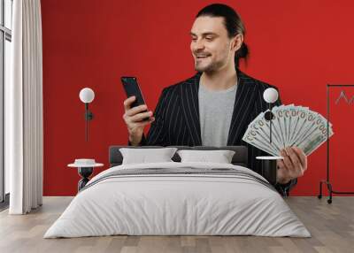 Young happy smiling successful rich latin man in black striped jacket grey shirt using mobile cell phone holding fan of cash money in dollar banknotes isolated on red color background studio portrait. Wall mural