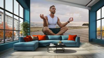 Young happy strong sporty athletic fit sportsman man wear sports clothes hold hands in yoga om aum gesture warm up training at sunrise sun over sea beach outdoor on pier seaside in summer day morning. Wall mural