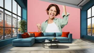 Young happy woman 20s she wear green shirt white t-shirt doing winner gesture celebrate clenching fists say yes isolated on plain pastel light pink background studio portrait People lifestyle concept Wall mural