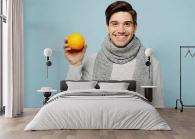 Young ill sick man wear gray sweater scarf hold in hand citrus orange fruit isolated on plain blue background studio portrait. Healthy lifestyle disease virus treatment cold season recovery concept. Wall mural