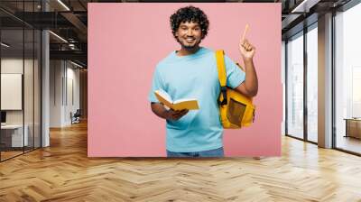 Young Indian boy student wear blue t-shirt casual clothes backpack bag write down in exercise book notebook point finger up isolated on plain pink background. High school university college concept. Wall mural