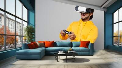 Young Indian man 20s he wearing casual yellow hoody hold in hand play pc game with joystick console watching in vr headset pc gadget isolated on plain grey background studio People lifestyle portrait Wall mural