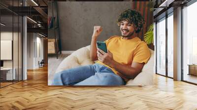 Young Indian man wear orange casual clothes hold use mobile cell phone do winner gesture sits in armchair stay at home hotel flat rest relax spend free spare time in living room indoor Lounge concept Wall mural