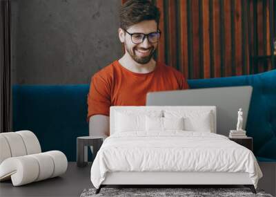 Young IT man wear red t-shirt glasses hold use work laptop pc computer sit on blue sofa couch stay at home flat rest relax spend free spare time in living room indoors grey wall People lounge concept. Wall mural