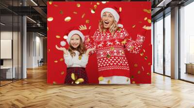 Young joyful woman in sweater hat have fun with child baby girl 6-7 years old. Mommy little kid daughter toss up confetti isolated on plain red color background studio. New Year love family concept. Wall mural