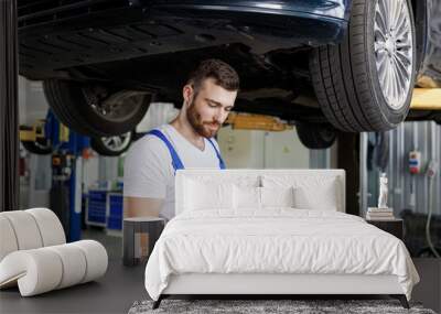 Young male professional technician mechanic man wears denim blue overalls t-shirt use hold mobile cell phone stand near car lift check technical condition work in vehicle repair shop workshop indoor Wall mural