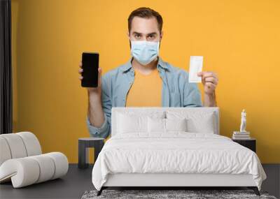 Young man in sterile face mask isolated on yellow background. Epidemic pandemic coronavirus 2019-ncov sars covid-19 flu virus concept Hold mobile phone with blank empty screen antiseptic alcohol wipe. Wall mural