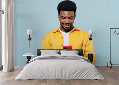 Young man of African American ethnicity 20s in yellow shirt hold in hand use mobile cell phone chatting online isolated on plain pastel light blue background studio portrait. People lifestyle concept. Wall mural