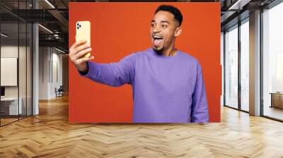 Young man of African American ethnicity in purple sweatshirt casual clothes do selfie shot on mobile cell phone post photo on social network isolated on plain red orange background Lifestyle concept Wall mural