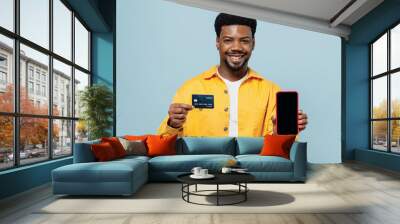 Young man of African American ethnicity in yellow shirt use mobile cell phone black screen workspace area credit bank card shopping online order delivery isolated on plain pastel light blue background Wall mural