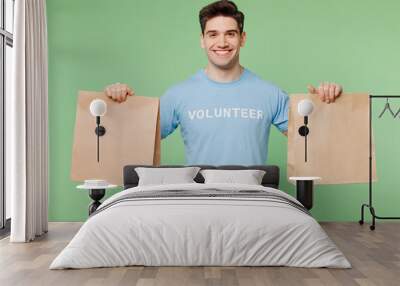 Young man wears blue t-shirt white title volunteer hold in hand two paper brown blank bags delivery isolated on plain pastel green background Voluntary free work assistance help charity grace concept Wall mural