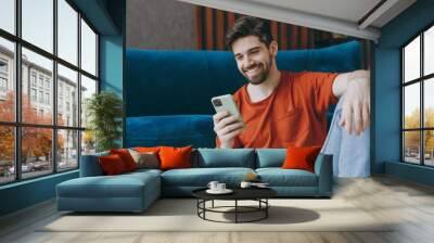 Young man wears red t-shirt hold in hand use mobile cell phone sit on blue sofa couch stay at home hotel flat rest relax spend free spare time in living room indoors grey wall. People lounge concept. Wall mural