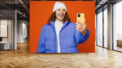 Young man with long curly hair wearing hat purple ski padded jacket casual clothes doing selfie shot on mobile cell phone post photo on social network isolated on plain orange red background studio. Wall mural