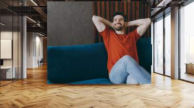 Young minded man wears red t-shirt hold hands behind neck look aside sit on blue sofa stay at home hotel flat rest relax spend free spare time in living room indoors grey wall. People lounge concept. Wall mural