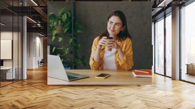 Young minded pensive employee business woman in casual yellow shirt hold takeaway delivery craft paper brown cup coffee to go sit work at wooden office desk with pc laptop Achievement career concept Wall mural