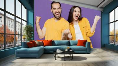 Young overjoyed happy couple two friends family man woman together in yellow casual clothes doing winner gesture celebrate clenching fists say yes isolated on plain violet background studio portrait. Wall mural