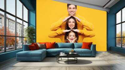 Young parents mom dad with child kid girl 7-8 years old wear pink sweater casual clothes stand behind each other hold hands above head pov roof isolated on plain yellow background. Family day concept. Wall mural