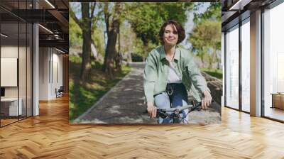 Young pensive dreamful happy woman 20s wearing casual green jacket jeans riding bicycle bike on sidewalk in city spring park outdoors, look aside People active urban healthy lifestyle cycling concept Wall mural