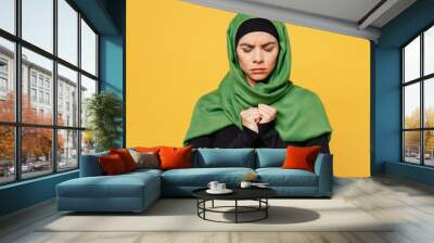 Young sad upset arabian asian muslim woman wearing green hijab abaya black clothes hold hand fists folded pray isolated on plain yellow background. People uae middle eastern islam religious concept. Wall mural