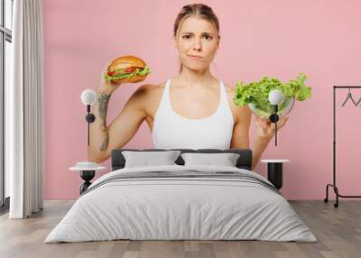 Young sad upset woman she wear white top casual clothes hold eat choose between lettuce salad and burger isolated on plain pink background. Proper nutrition healthy fast food unhealthy choice concept. Wall mural