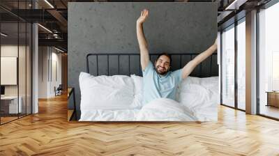 Young satisfied smilng happy fun man wear casual blue t-shirt lying in bed stretch hands rest relax spend time in bedroom lounge home in own room house wake up dream. Good mood night bedtime concept. Wall mural