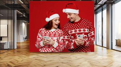 Young shocked couple friends two man woman in sweater hat hold in hand use mobile cell phone isolated on plain red background studio. Happy New Year 2022 celebration merry ho x-mas holiday concept Wall mural