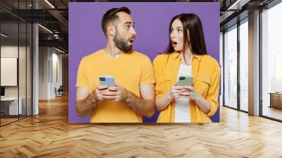 Young shocked surprised couple two friends family man woman together in yellow casual clothes hold in hand use mobile cell phone chatting look to each other isolated on plain violet background studio. Wall mural