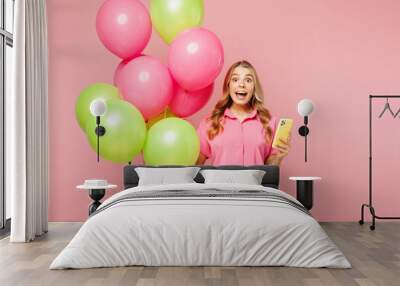 Young shocked woman wear shirt celebrating birthday holiday and hold bunch of colorful air balloons hold in hand use mobile cell phone isolated on plain pastel light pink background Lifestyle concept Wall mural