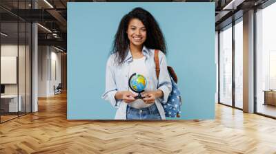 Young smiling african american girl teen geography student wear denim clothes backpack hold in hands Earth world globe isolated on blue background Education in high school university college concept. Wall mural