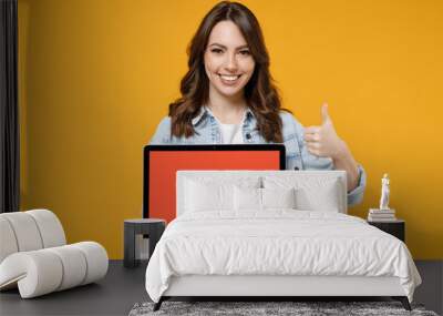 Young smiling brunette fun woman 20s wear denim shirt white t-shirt hold laptop pc computer with blank screen workspace area show thumb up like gesture isolated on yellow background studio portrait Wall mural