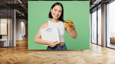 Young smiling cheerful fun woman wearing white clothes eat burger hold scales look camera isolated on plain pastel light green background. Proper nutrition healthy fast food unhealthy choice concept. Wall mural