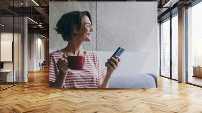 Young smiling dreamful calm happy 20s wear casual clothes sitting on grey sofa using mobile cell phone hold red cup drink coffee look aside rest indoors flat at home People lifestyle leisure concept Wall mural