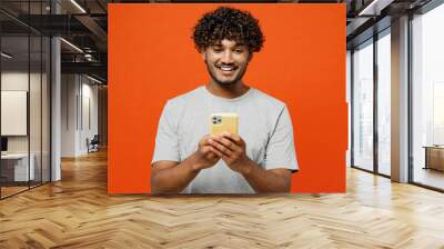 Young smiling fun cool happy Indian man he wears t-shirt casual clothes hold in hand use mobile cell phone browsing internet isolated on orange red color background studio portrait. Lifestyle concept. Wall mural