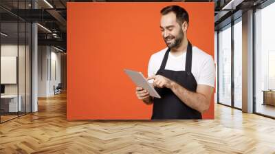 Young smiling fun man 20s barista bartender barman employee in black apron white t-shirt work in coffee shop using tablet pc computer browsing internet isolated on orange background. business startup. Wall mural