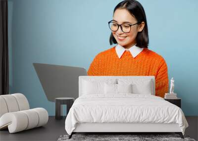 Young smiling fun smart IT woman of Asian ethnicity wear orange sweater glasses hold use work on laptop pc computer isolated on plain pastel light blue cyan background studio People lifestyle concept Wall mural