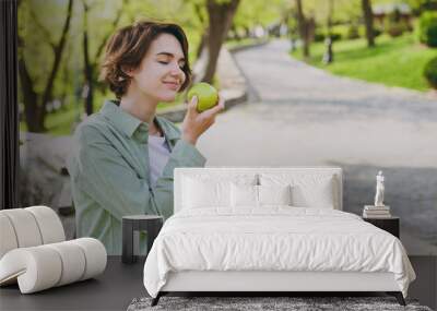 Young smiling happy caucasian student woman 20s in casual green jacket jeans sitting on bench in city spring park outdoors resting eat apple fruit. People vegeterian healthy urban lifestyle concept Wall mural