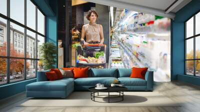 Young smiling happy caucasian woman 20s wear casual clothes shopping at supermaket store with grocery cart buy choose milk dairy produce inside hypermarket. People purchasing gastronomy food concept Wall mural