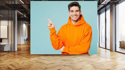 Young smiling happy fun man he wears orange hoody casual clothes point index finger aside on area mock up isolated on plain pastel light blue cyan color background studio portrait. Lifestyle concept. Wall mural