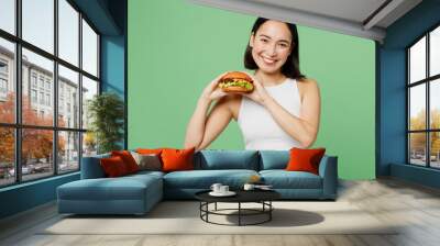 Young smiling happy fun woman wearing white clothes holding eating burger looking camera isolated on plain pastel light green background. Proper nutrition healthy fast food unhealthy choice concept. Wall mural