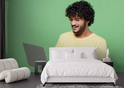 Young smiling happy Indian man 20s in basic yellow t-shirt hold use work on laptop pc computer chatting online isolated on plain pastel light green background studio portrait People lifestyle concept Wall mural