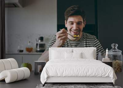 Young smiling happy man in casual clothes striped t-shirt eat breakfast muesli cereals with milk fruit in bowl sit by table cooking food in light kitchen at home alone Healthy diet lifestyle concept. Wall mural