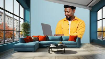 Young smiling happy man of African American ethnicity 20s wear yellow shirt hold use work on laptop pc computer isolated on plain pastel light blue background studio portrait People lifestyle concept Wall mural