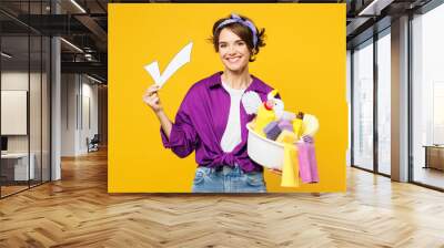 Young smiling happy woman wear purple shirt rubber gloves hold basin with detergent bottles do housework tidy up hold in hand check mark sign isolated on plain yellow background. Housekeeping concept. Wall mural