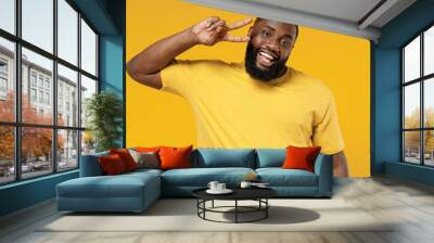 Young smiling satisfied cheerful fun happy black man 20s wearing bright casual t-shirt cover eye with victory sign isolated on plain yellow color background studio portrait. People lifestyle concept Wall mural