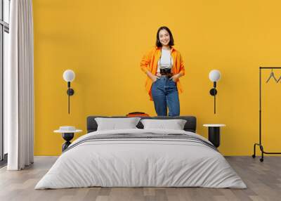 Young smiling woman wear summer casual clothes stand near suitcase baggage isolated on plain yellow background. Tourist travel abroad in free spare time rest getaway. Air flight trip journey concept. Wall mural